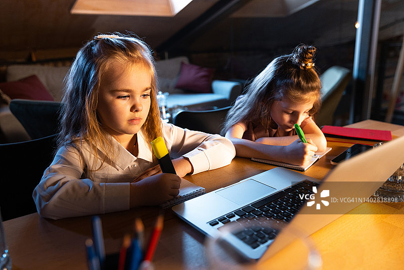 两个小可爱的孩子，姐妹坐在家里，晚上在家做作业。在线教育、童年、家庭、远程学习和学校理念。图片素材