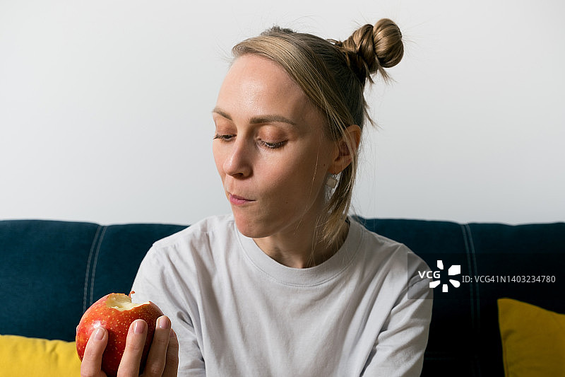 一位坐在色彩鲜艳的沙发上的女士手里拿着一个红苹果。女孩吃了一个水果，咬下一块多汁的苹果。种植有机农产品。素食的概念，纯素，生食和饮食。图片素材