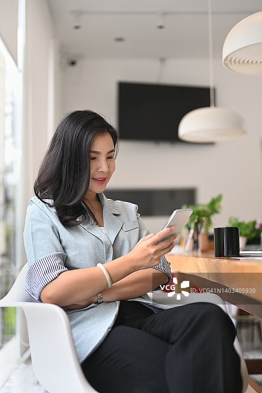 漂亮年轻的亚洲女性使用智能手机，查看社交媒体，短信按摩，而坐在现代工作场所和使用智能手机。图片素材