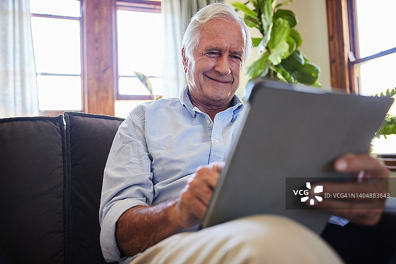 微笑的老人在家里的沙发上使用平板电脑图片素材