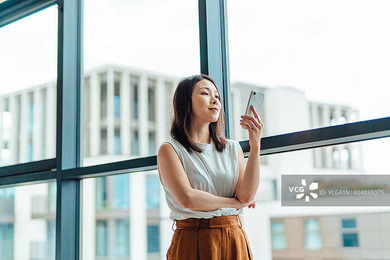 用智能手机的年轻商务女性站在办公室窗口的低角度视角图片素材