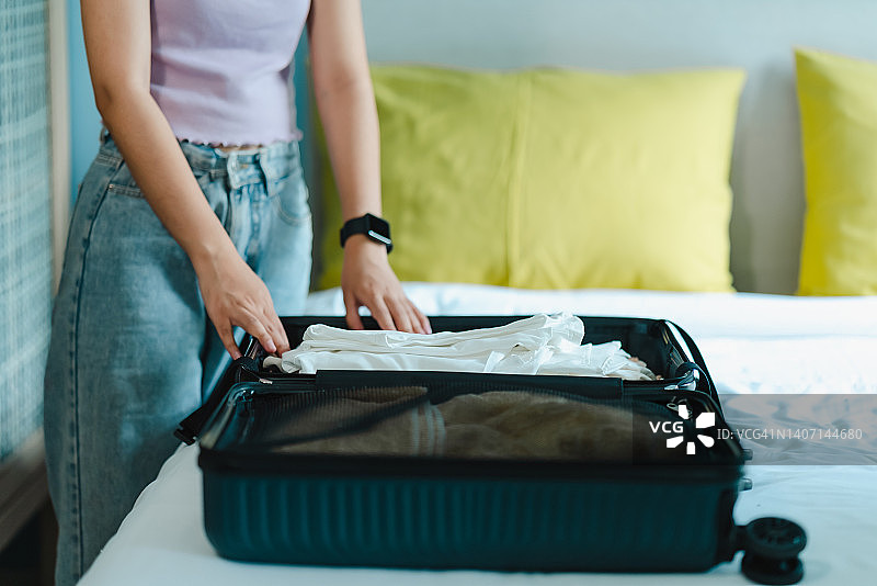 一位留着长发的亚洲少女坐在卧室的床上，把衣服叠在一个大行李箱里，为周末旅行做准备。打包行李，在出发前检查是否有遗忘物品。图片素材