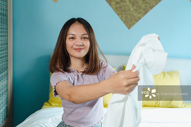 一个十几岁的亚洲妇女检查一件白色衬衫从一个大手提箱。为了周末旅行，表现得像个新手游客。一位女士到达酒店后正在拿出衣服试穿。图片素材