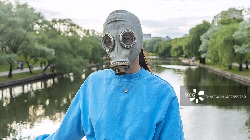 一个戴防毒面具的女孩站在河上的一座桥上。城市中的疏离感。时尚、生态、孤独的概念。图片素材