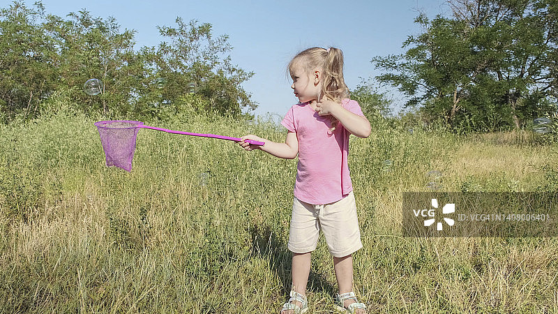 小女孩在公园的高草丛中玩耍，用蝴蝶网捕捉肥皂泡。可爱的小女孩在阳光灿烂的日子里在草地上用空中捕虫网捉肥皂泡。图片素材