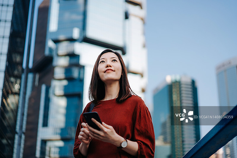 自信而成功的年轻亚洲女商人在金融区使用智能手机，对抗城市的摩天大楼。坚定的女性企业家，用智能手机仰望天空。工作在进行中。职场中的女性领导图片素材