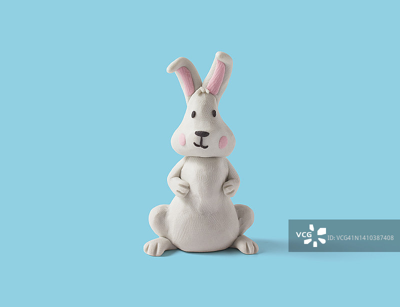蓝色背景的白色橡皮泥做的可爱坐兔。手工制作的粘土复活节兔子。三维作品图片素材