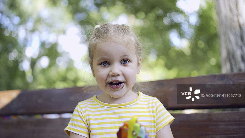 小女孩拿着一个五颜六色的姜饼微笑着。特写可爱的小女孩坐在公园长凳上饼干看着相机镜头。图片素材
