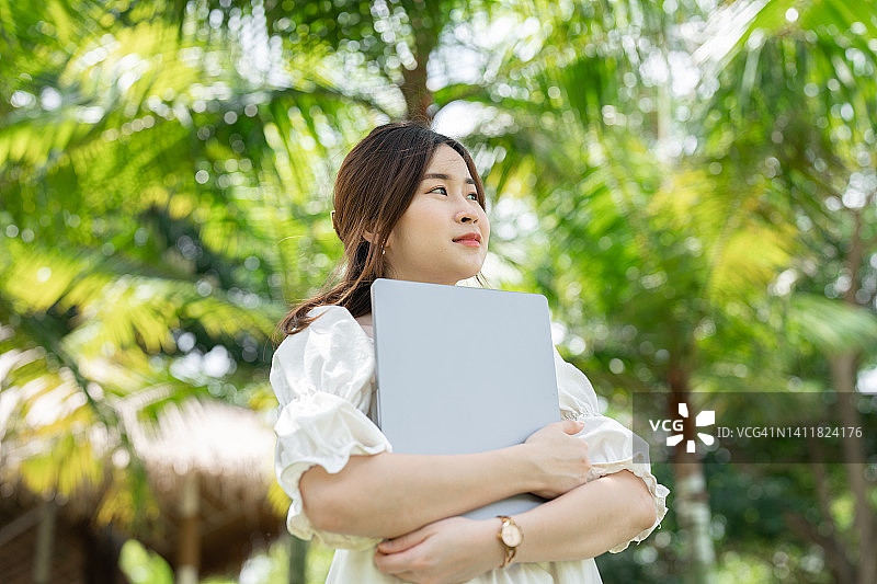 自信和成功的年轻亚洲女商人的肖像手持笔记本电脑对绿色的自然植物。忙碌的工作图片素材