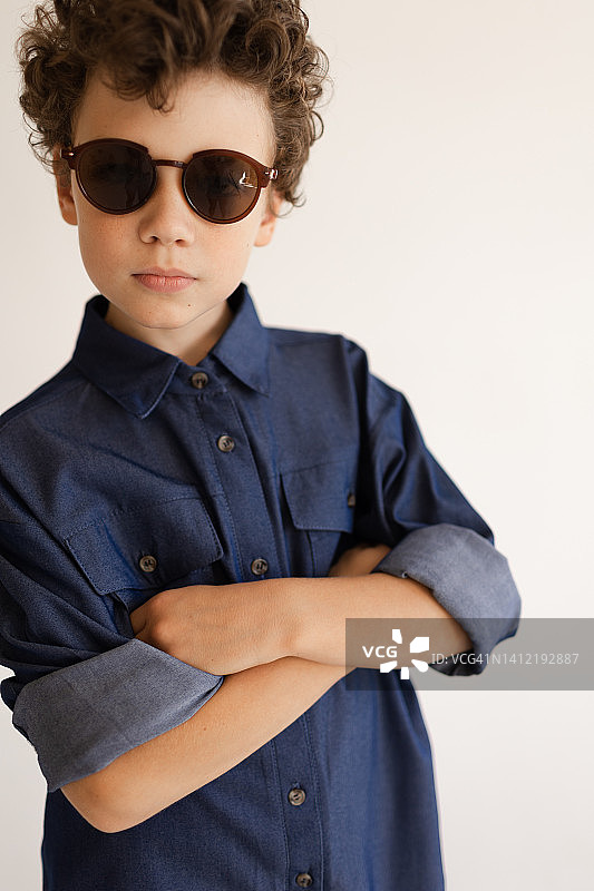 一个卷发男孩的肖像，戴着太阳镜，牛仔衬衫和牛仔裤在一个白色的背景上的工作室图片素材
