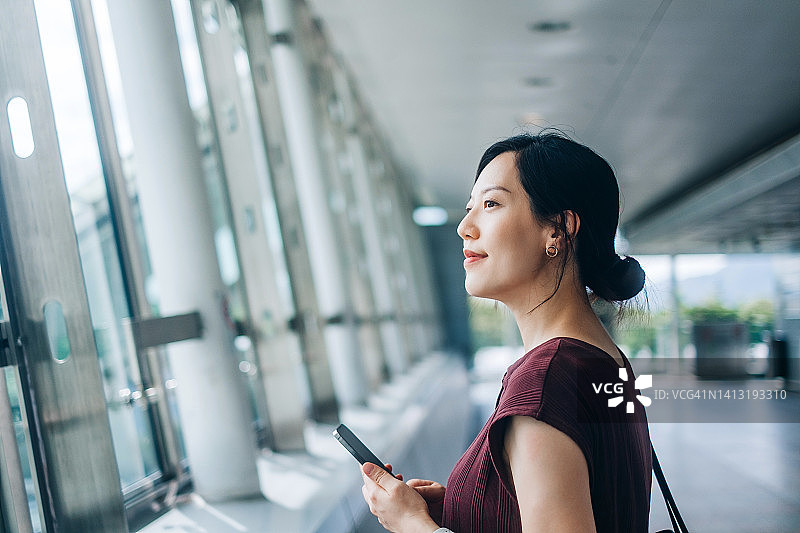 自信的年轻亚洲女商人拿着智能手机看向窗外。一位成功的女性高管站在一个现代化的办公空间里，靠窗眺望前方。女性领导力。忙碌的工作图片素材