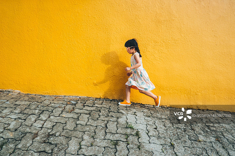 可爱的亚洲小女孩的侧面，戴着墨镜走在人行道上对着黄色的墙图片素材