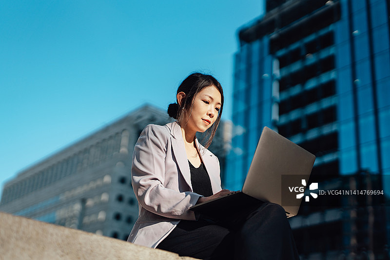 年轻的女商人坐在金融大楼旁，用笔记本电脑工作图片素材