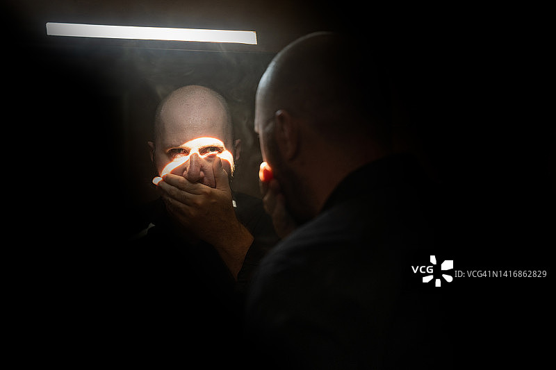 一个严肃的人用手闭上嘴，站在黑暗的房间里的镜子前，透过镜子看着相机图片素材