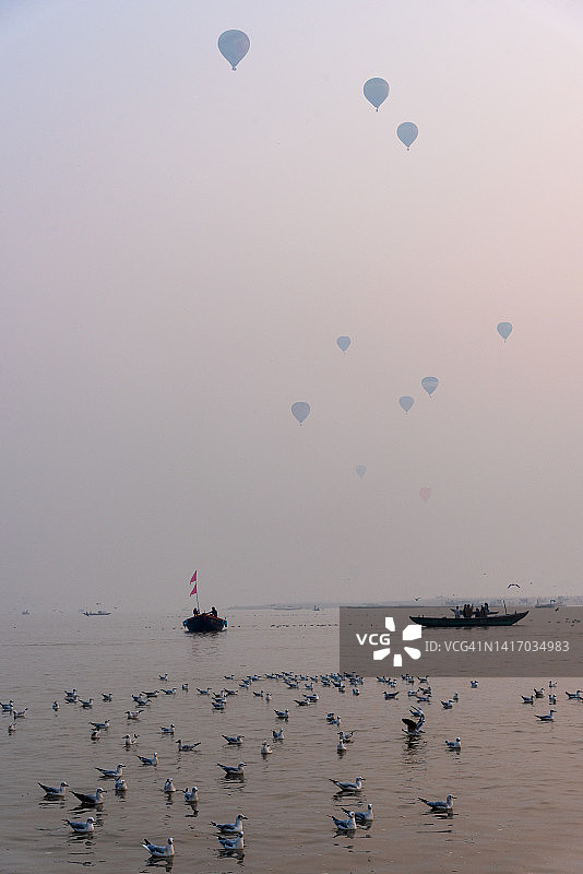 在印度北方邦瓦拉纳西的排灯节期间，恒河上的船只和鸟儿在早晨与热气球作背景。图片素材