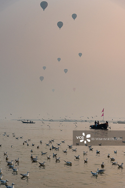 在印度北方邦瓦拉纳西的排灯节期间，恒河上的船只和鸟儿在早晨与热气球作背景。图片素材