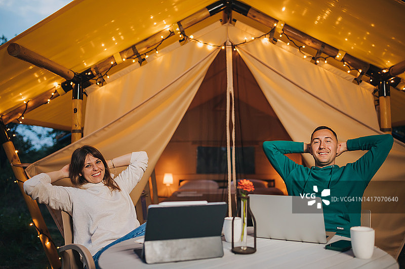 夏日傍晚，一对幸福的自由家庭夫妇在舒适的野营帐篷里用笔记本电脑工作。用于户外度假和度假的豪华露营帐篷。生活方式的概念图片素材
