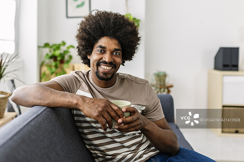 微笑肖像的年轻非洲裔美国人放松在沙发上喝咖啡图片素材