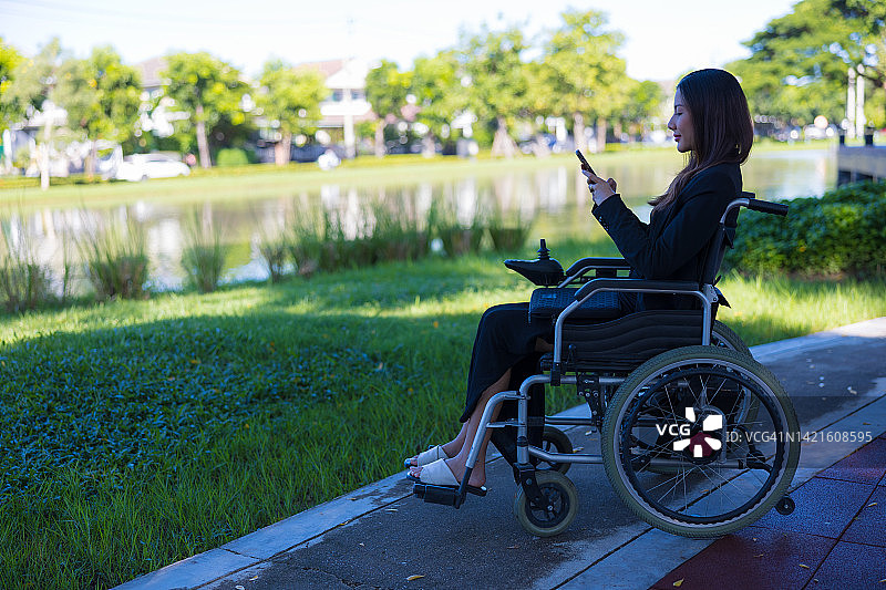 亚洲女性坐在机动轮椅上，在公园里打电话。图片素材