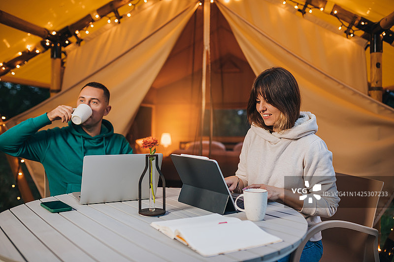 幸福的家庭夫妇自由职业者在一个舒适的野营帐篷在夏天的晚上使用笔记本电脑。户外度假度假的豪华露营帐篷。生活方式的概念图片素材