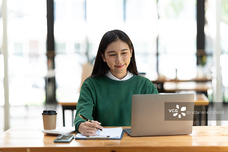 年轻的女人在办公室的笔记本电脑工作，快乐的年轻亚洲女商人坐在她的工作场所在办公室。图片素材