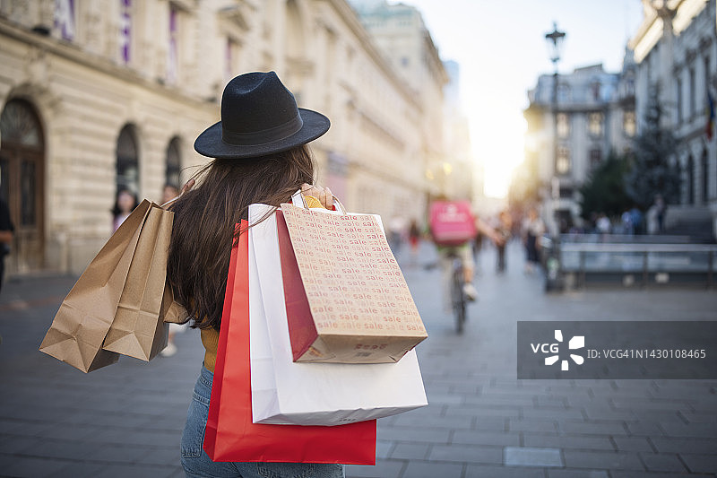 背面的女人戴着帽子走在城市的街道上，拿着购物袋。图片素材