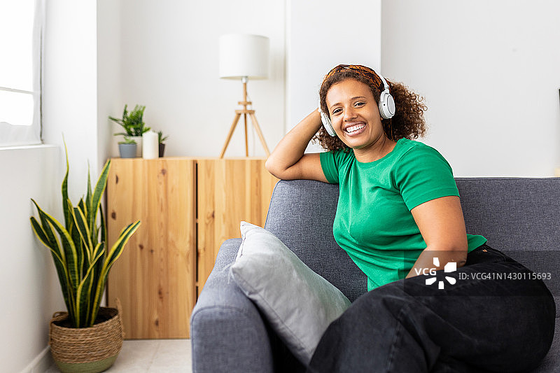 快乐的千年非洲妇女放松在沙发上听音乐对着镜头微笑图片素材