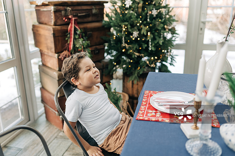 快乐的非裔美国男孩戴着圣诞帽，坐在桌子旁，看着镜头。一家人在家里共度圣诞佳节。图片素材