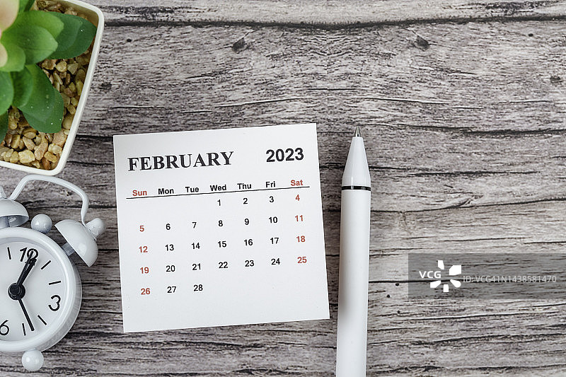 日历桌2023:2月是组织者计划和截止日期的月份，木桌背景上有一个闹钟和白笔。图片素材
