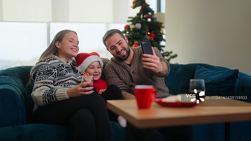 圣诞节期间，年轻的家庭坐在家里的客厅里，通过视频电话与其他家庭成员或朋友联系图片素材
