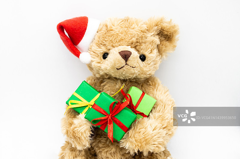 一只泰迪熊戴着红色的圣诞老人帽子，一只耳朵上戴着绒球，爪子里拿着绿色的礼盒。白色背景，复制空间图片素材