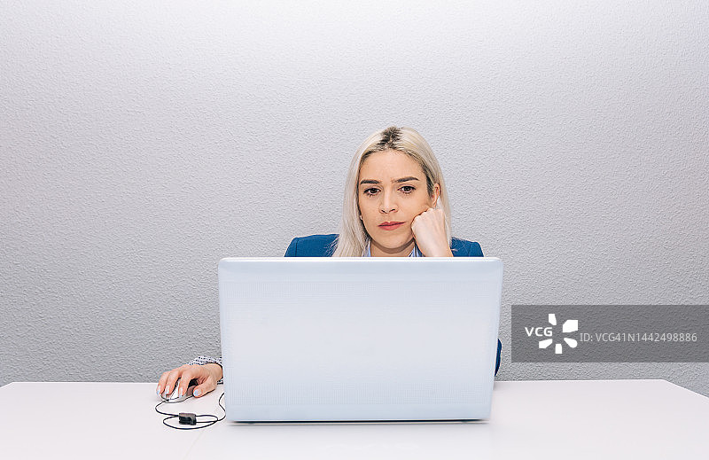 金发碧眼的年轻女子，身穿一件蓝色西装外套，用笔记本电脑在家远程办公。远程工作的概念。图片素材