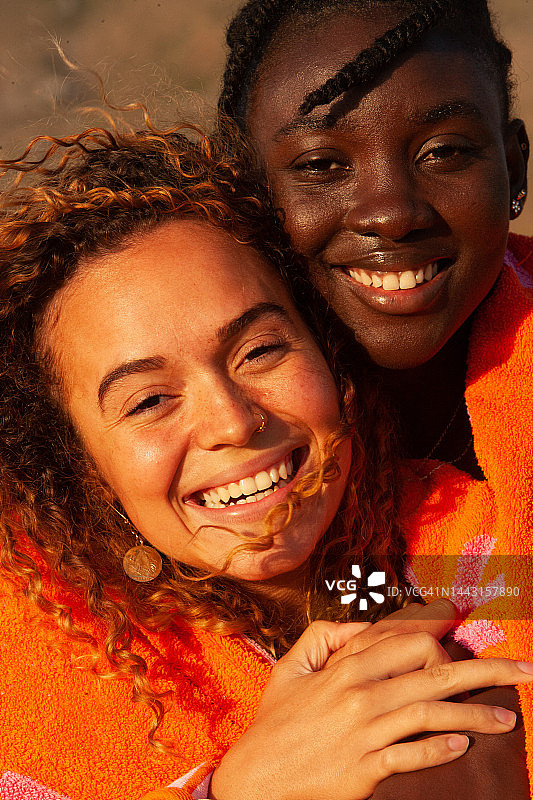 两个喜气洋洋的美丽年轻女子看着镜头，两个二十多岁的女孩和那里的女孩朋友与自然辫子和覆盖着橙粉色的毛巾是她的朋友长卷金色头发后面图片素材