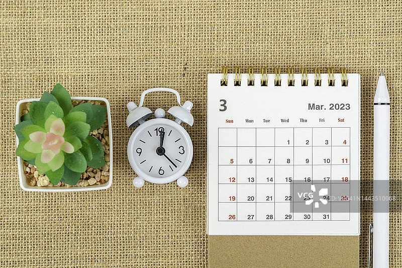 日历桌2023:3月是组织者制定计划和期限的月份，有闹钟、室内植物和粗麻布背景的白色钢笔。图片素材