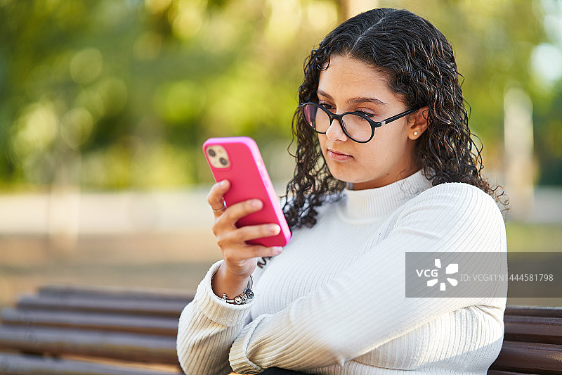 一名戴眼镜的妇女坐在公园的长凳上使用手机。图片素材