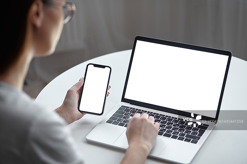 样机的白色屏幕笔记本电脑和手机妇女使用电脑，而坐在桌子在家里，背影图片素材
