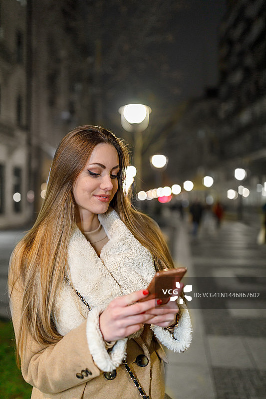 一个年轻的女人晚上走在城市的街道上用电话。图片素材