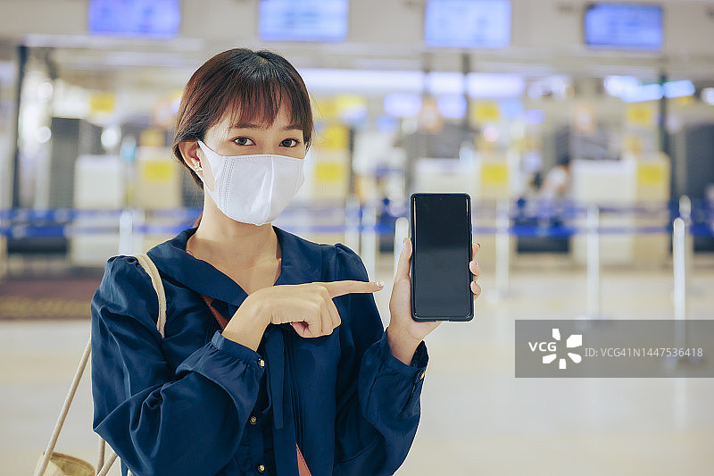 年轻的亚洲女商人戴着口罩，手持空屏手机在机场离境区。商务旅行的概念。图片素材