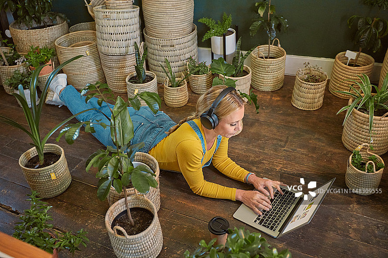 植物，苗圃和地板上的女人用笔记本电脑在线工作，使用互联网和流媒体音乐。园艺，室内园艺和女孩戴着耳机用电脑听互联网音频图片素材