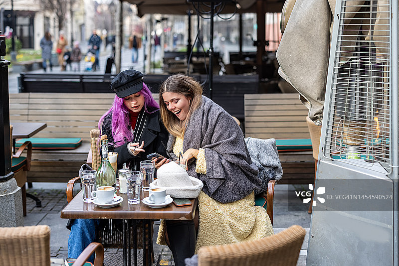 两个穿着暖和衣服的年轻女子坐在当地的咖啡馆里看手机。图片素材
