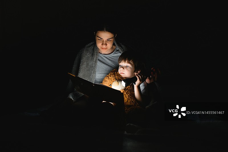 幸福的家庭。是时候讲故事了。快乐可爱的妈妈和儿子在睡觉前享受一本书，并使用手电筒。图片素材