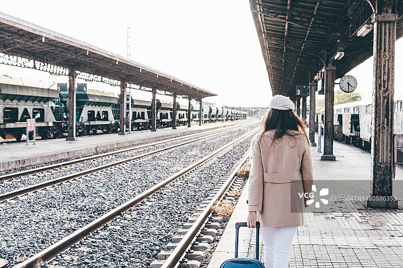 一个女孩戴着贝雷帽，穿着米色夹克，提着行李箱穿过火车站。图片素材