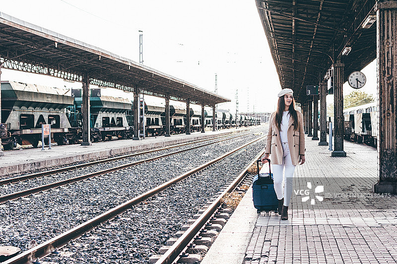 一个戴着贝雷帽和米色夹克的女人的肖像，她带着行李箱穿过火车站。图片素材