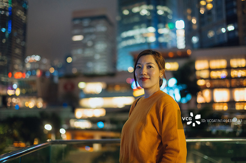 微笑的年轻亚洲女商人站在繁忙的市中心的建筑露台上的肖像。自信的职业女性在城市里，看着镜头，以五彩缤纷繁华的城市夜景为背景图片素材