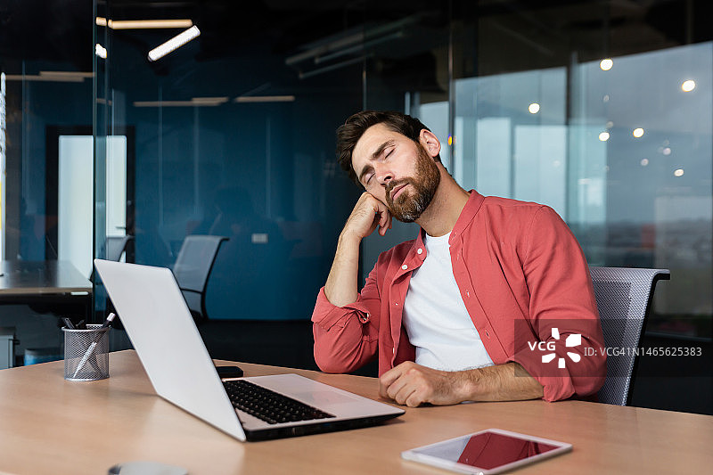 穿着休闲红衬衫的疲惫商人在工作场所睡觉，办公桌上的人在办公时间用笔记本电脑睡着了图片素材