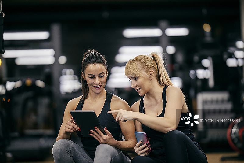 在健身房里，一位快乐的女运动员正指着平板电脑，她的女私人教练正在展示她的进步。图片素材