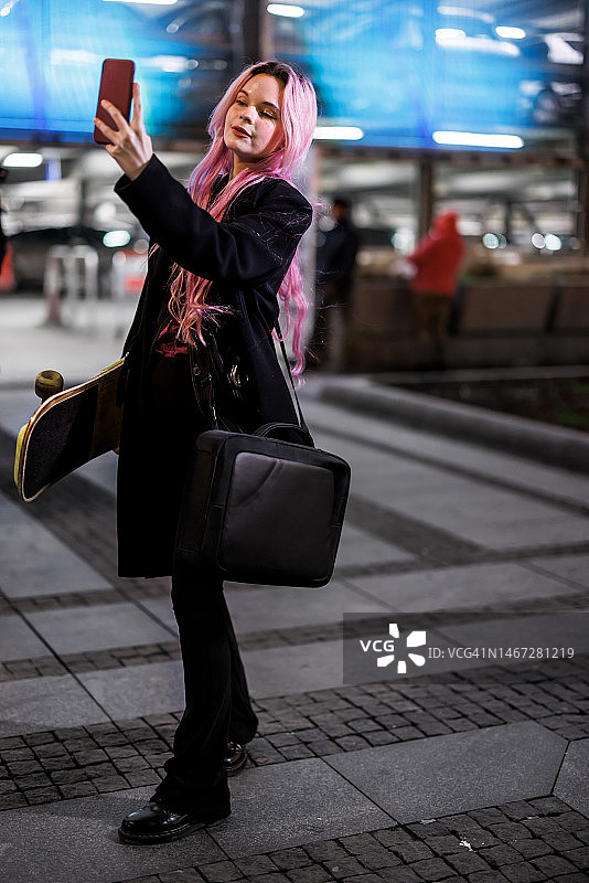 一个美丽的女人在城市里站着滑板，用她的手机自拍。图片素材