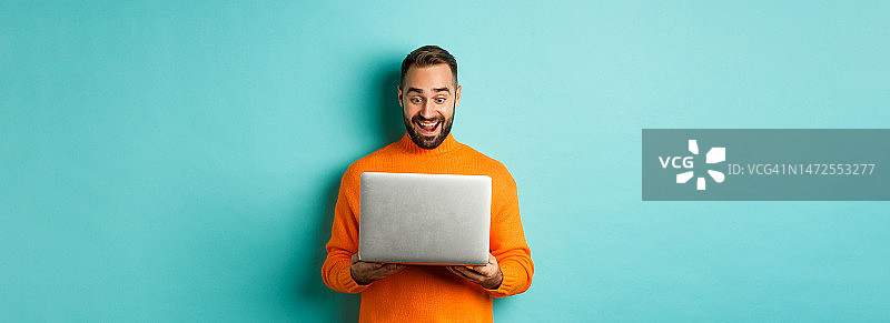 穿着橙色毛衣的兴奋男子在笔记本电脑上工作，看着电脑图片素材
