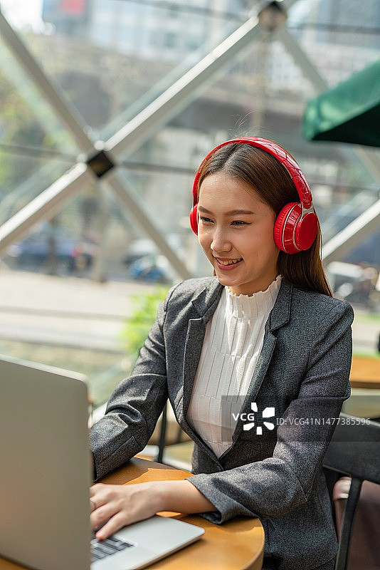 混合式工作风格。年轻的亚洲女性成年人与笔记本电脑在咖啡馆工作。戴着红色耳机的女商人图片素材