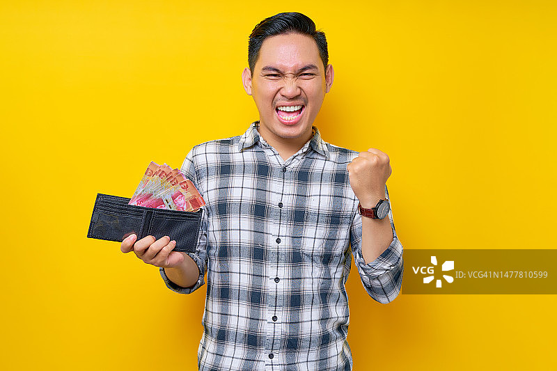 兴奋的年轻亚洲男子的肖像，穿着格子衬衫，手里拿着一个装满现金的钱包印尼盾钞票，庆祝成功孤立在黄色背景。人们的生活方式理念图片素材